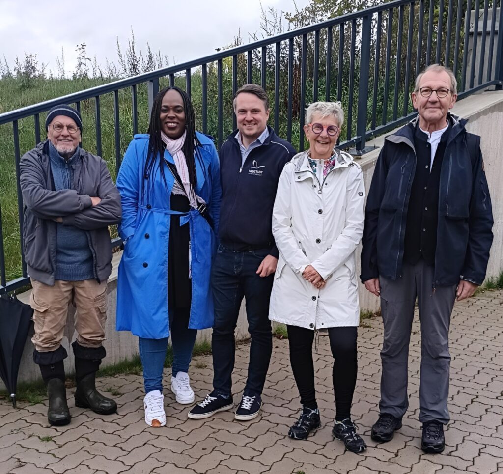 Fünf Personen, darunter der Bürgermeister von Neustadt und Vertreter*innen der Grünen, stehen lächelnd vor dem Deich; Geländer, Wiese und bewölkter Himmel im Hintergrund.